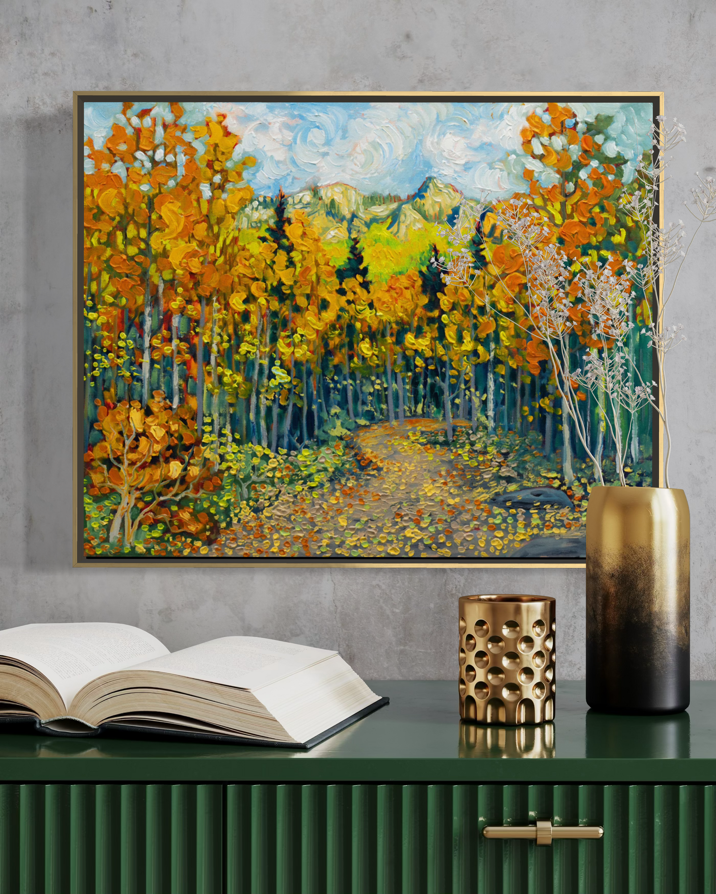 A framed oil painting of an autumn forest with golden yellow and orange trees, green mountains, and a blue sky, displayed on a dark green cabinet against a textured gray wall.